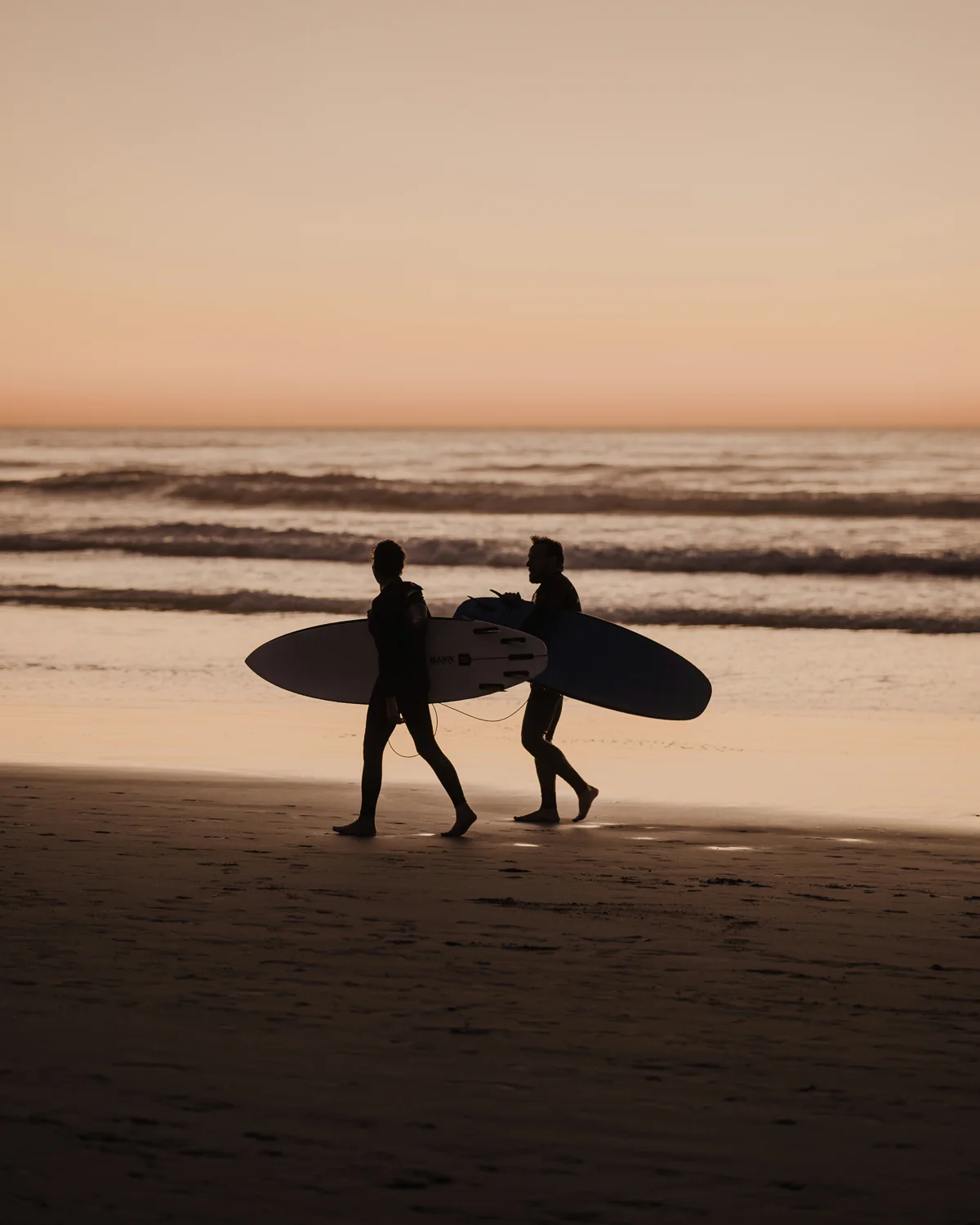 Two surfers walking the beach at sunset, boards under arm.