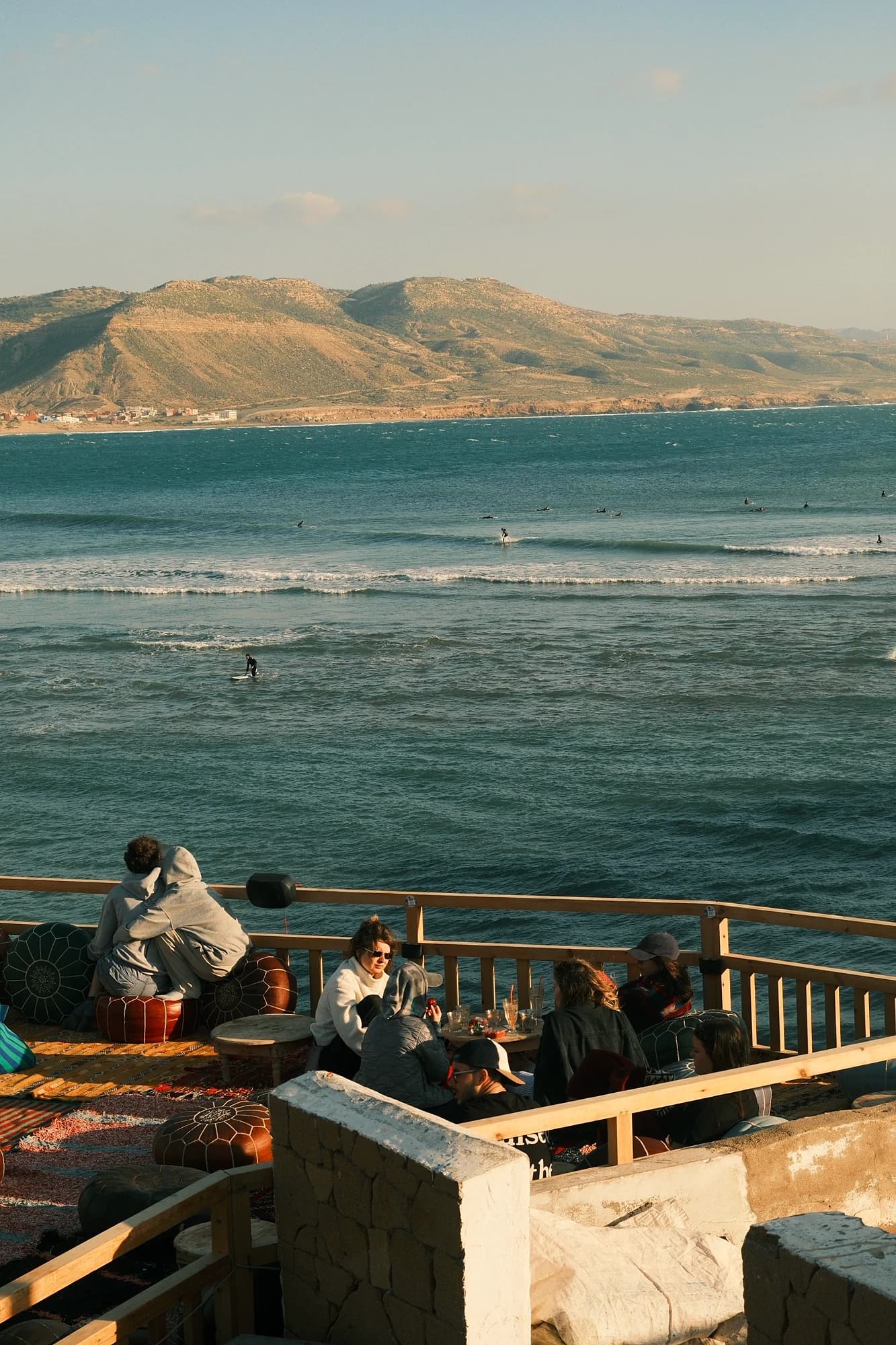 A terrace above Taghazout bay at golden hour, surfers in the lineup below